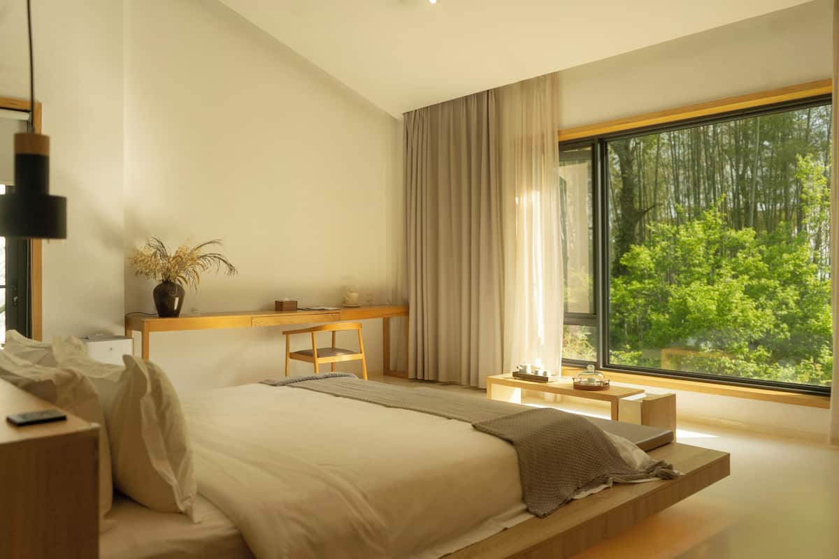A modern bedroom with a large bed, bedside tables, a desk, and a view of a forest through a large window.