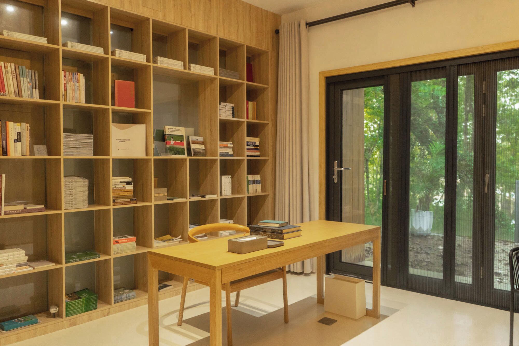 A room with a wooden bookshelf filled with books, a wooden table with a chair, and a sliding glass door leading to an outdoor area.