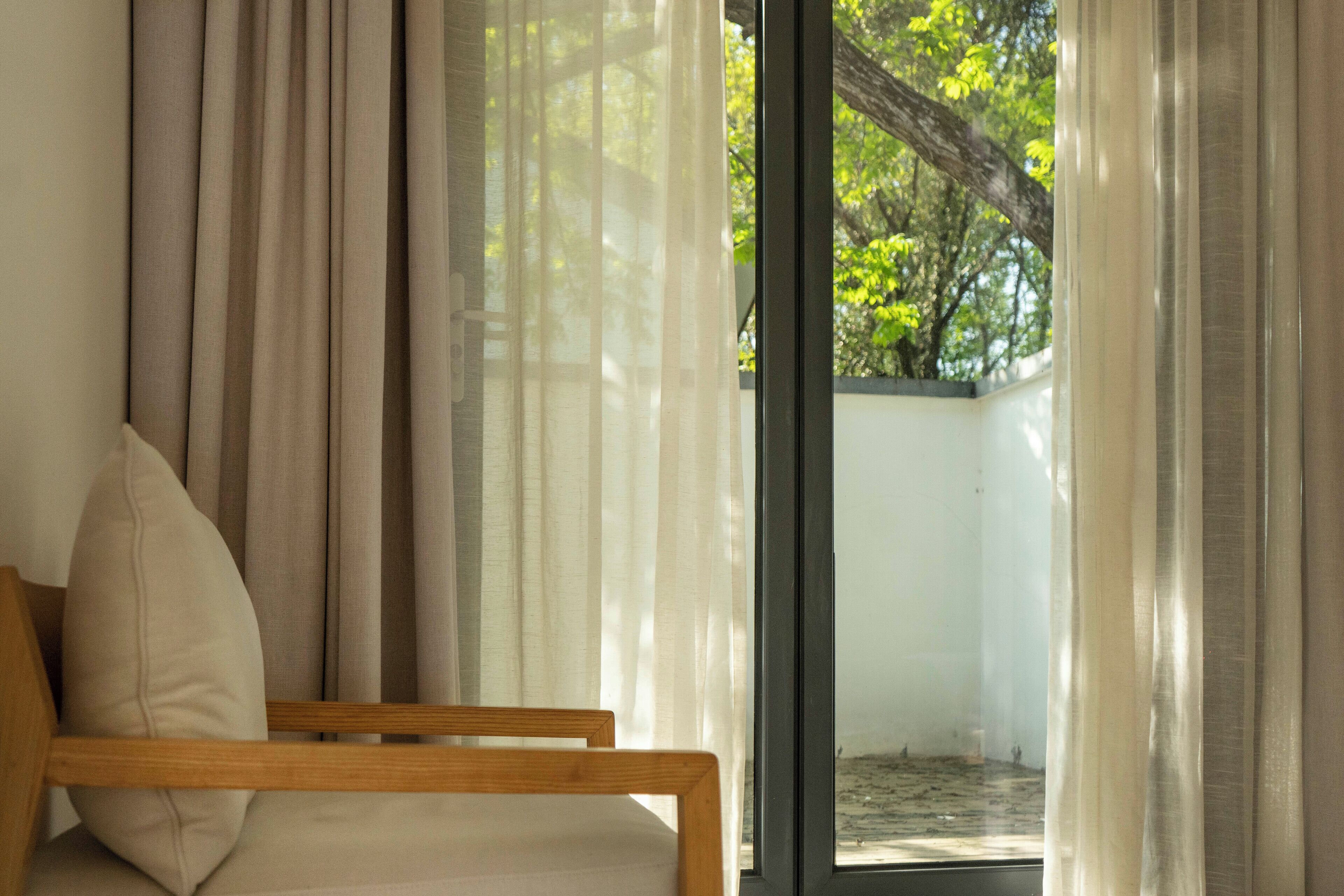 Una habitación con una silla de madera y vistas a través de una ventana con cortinas translúcidas.
