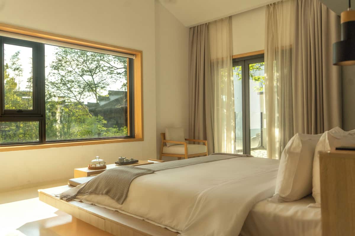A bedroom with a large window, a bed, a chair, and a bedside table.
