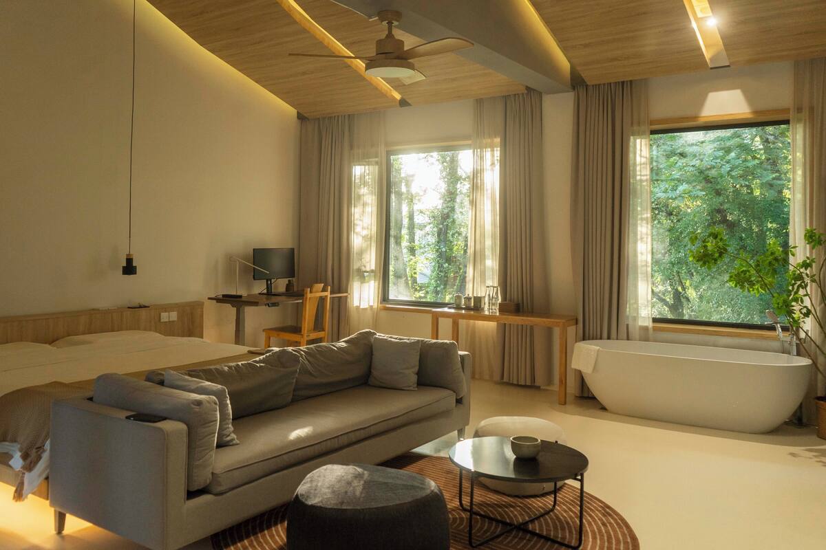 A modern hotel room with a bed, sofa, round coffee table, and a bathtub.