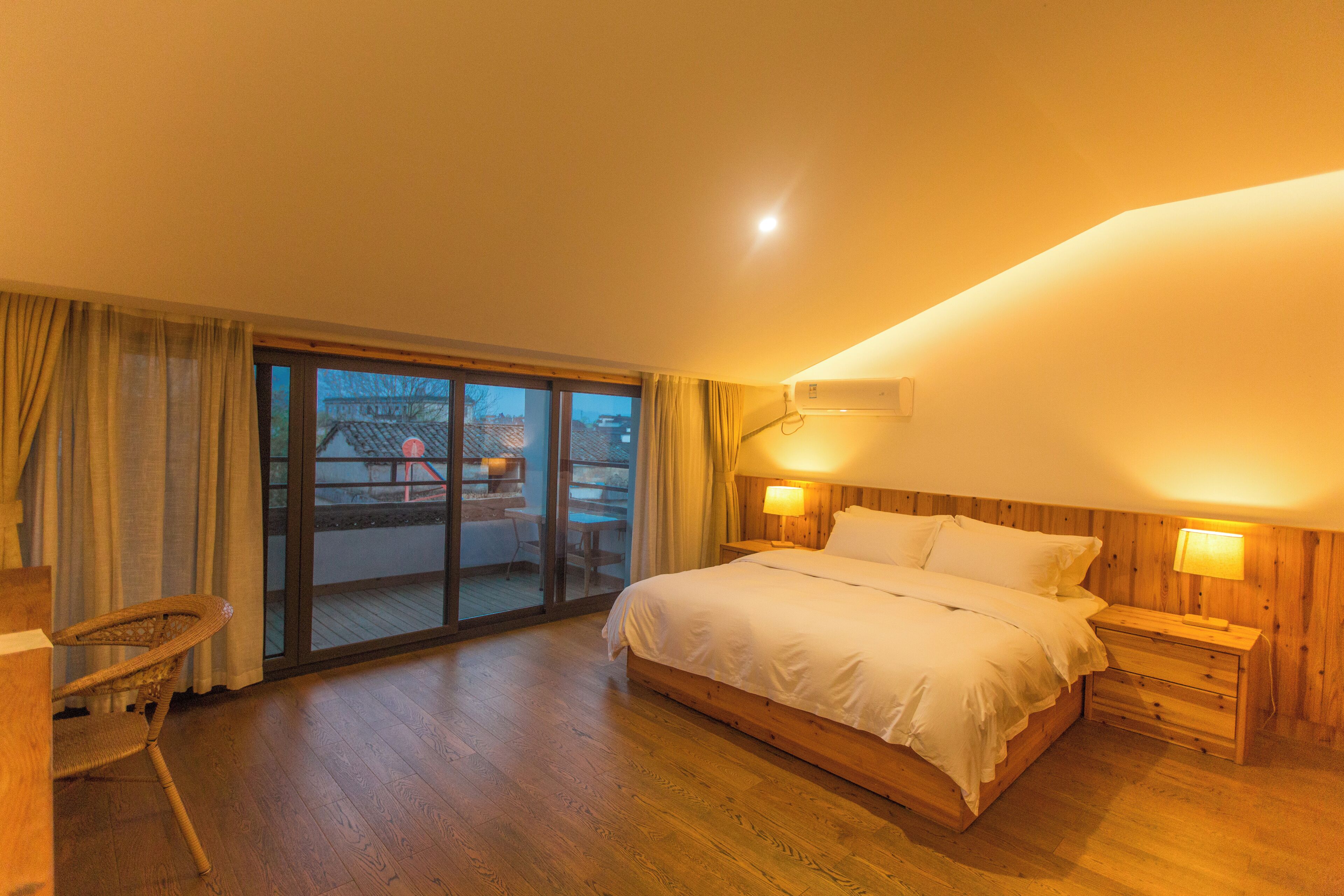 A bedroom with a large bed, wooden headboard, two bedside tables with lamps, a wooden floor, a sliding glass door leading to an outdoor area, and a wicker chair.