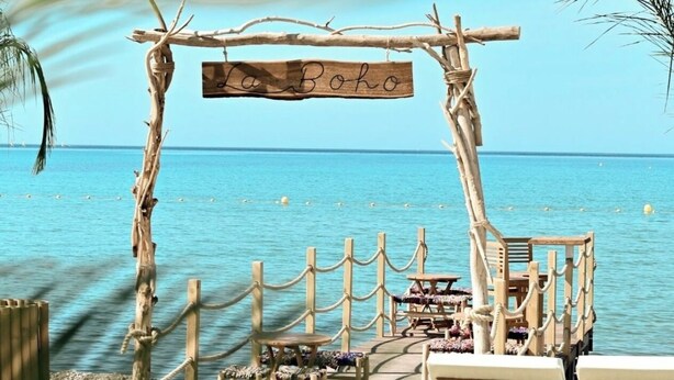 Eine Holzterrasse mit Liegestühlen und einem Schild mit der Aufschrift 'La Boho', mit Blick auf das klare, blaue Meer.