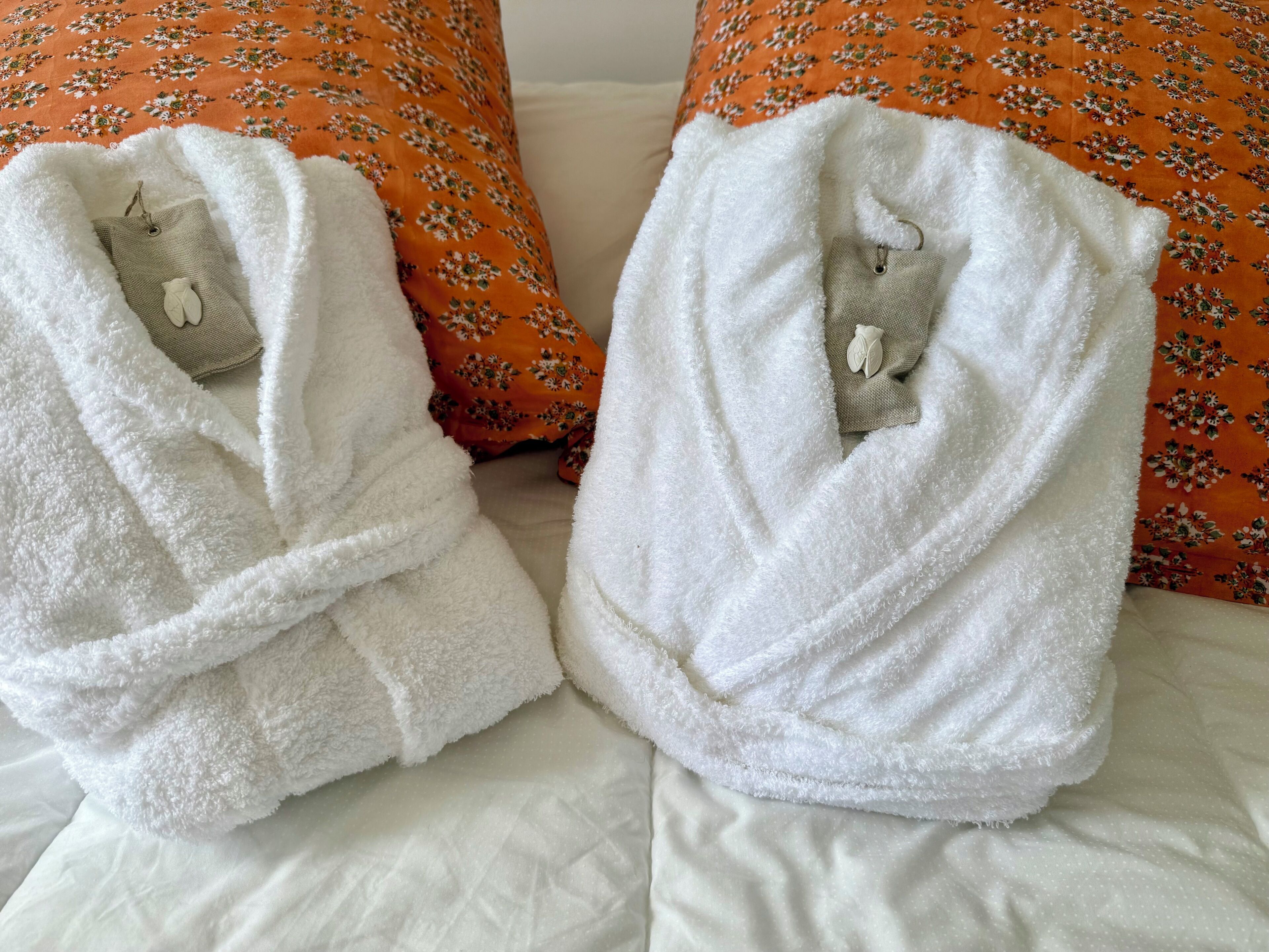 Dois roupões brancos sobre uma cama com um travesseiro floral laranja e um travesseiro branco.