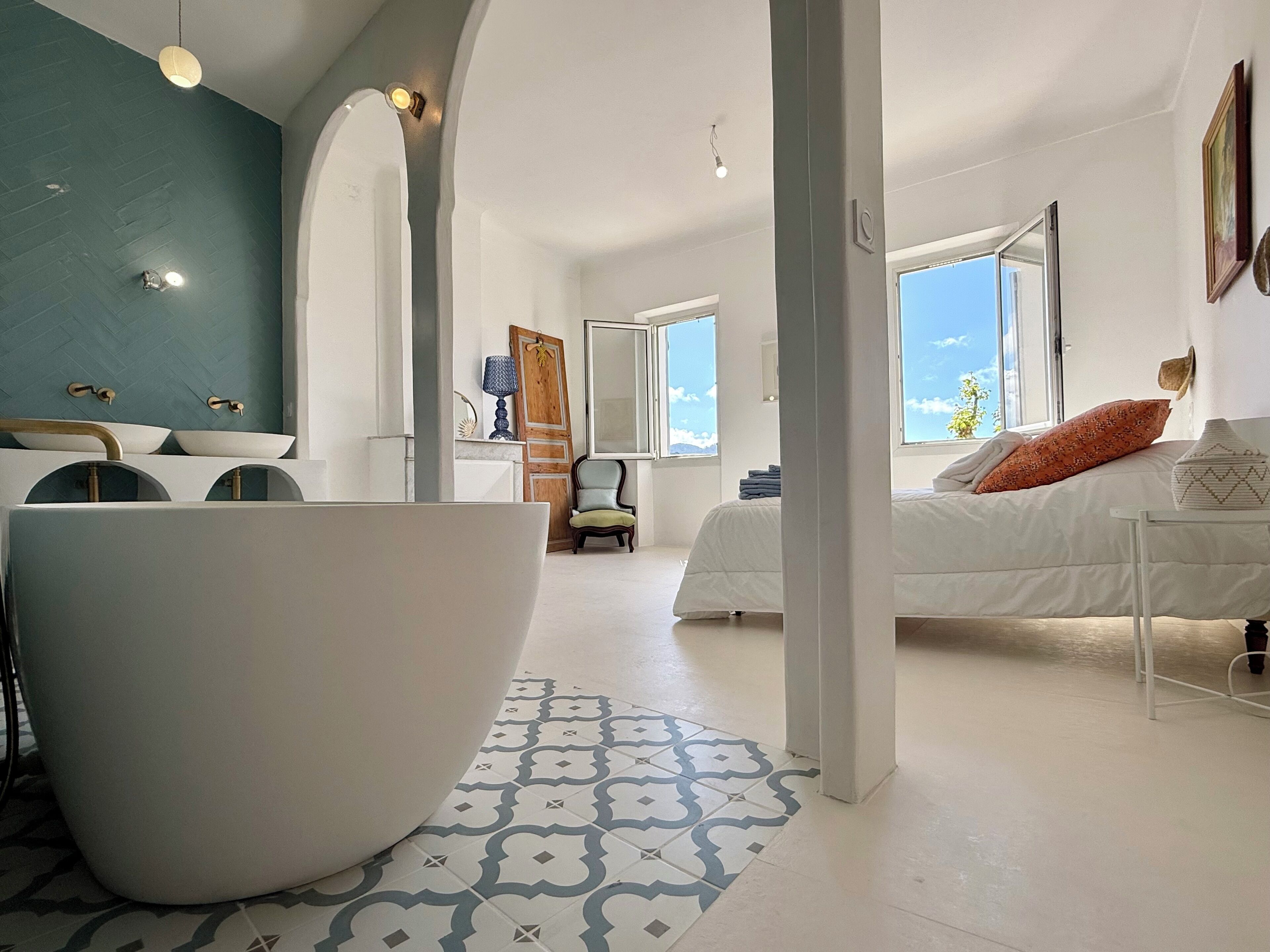 A modern hotel room with a large bathtub, a bed with a white duvet, a chair, and a large window offering a view of the sky.