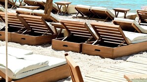 Ein Strand mit Holzliegen und kleinen Tischen, angeordnet auf sandigem Gelände mit Blick auf das Meer und eine entfernte Insel.