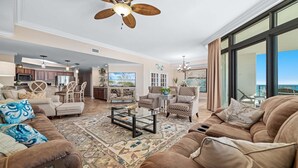 A spacious living room with a large sofa, armchairs, a coffee table, and a patterned rug. There is a ceiling fan, a kitchen in the background, and a view of the ocean through large windows. - Beachfront 3BR, Lazy River/Slide, On-site Cafe and Bar! (Orange Beach)