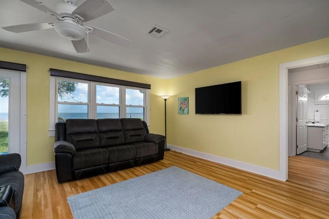 A room with a black leather sofa, a grey rug, a ceiling fan, a TV mounted on the wall, and a window with a view of the outdoors.