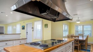 A kitchen with a central island, white cabinetry, a stainless steel range hood, and a dining area with a table and chairs. - Island Time 3 Bedroom Home (Rockport)