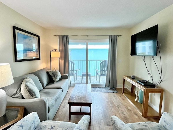Een woonkamer met een bank, fauteuils, een salontafel, een televisie en een balkon met stoelen.