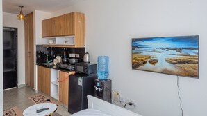 A compact kitchen with wooden cabinets, a refrigerator, a microwave, and a water dispenser. There's a bed with a white headboard and a painting of a landscape on the wall.