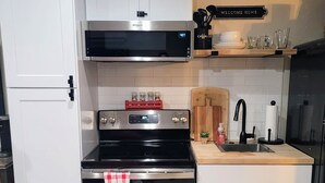 Una cocina moderna con armarios blancos, una campana extractora de acero inoxidable, horno empotrado y microondas. En la encimera hay una tabla de cortar y una máquina de café.