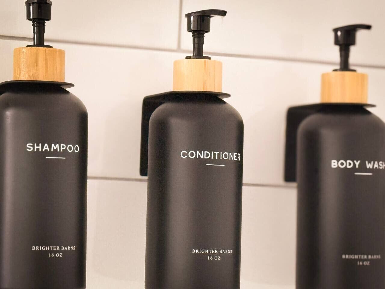 Drie flessen Brightener Barns producten: shampoo, conditioner en douchegel, elk met een zwarte verpakking en houten pomp.