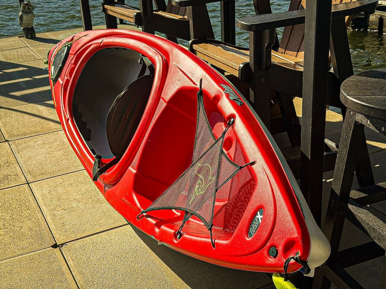 A red kayak with a netted cover, placed on a tiled surface near wooden chairs and tables.