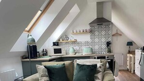 A loft-style living space with a high ceiling, a sofa with cushions, a striped ottoman, and a kitchen area with a microwave, kettle, and decorative items. - Grove View Studio Apartment. (Cossington)
