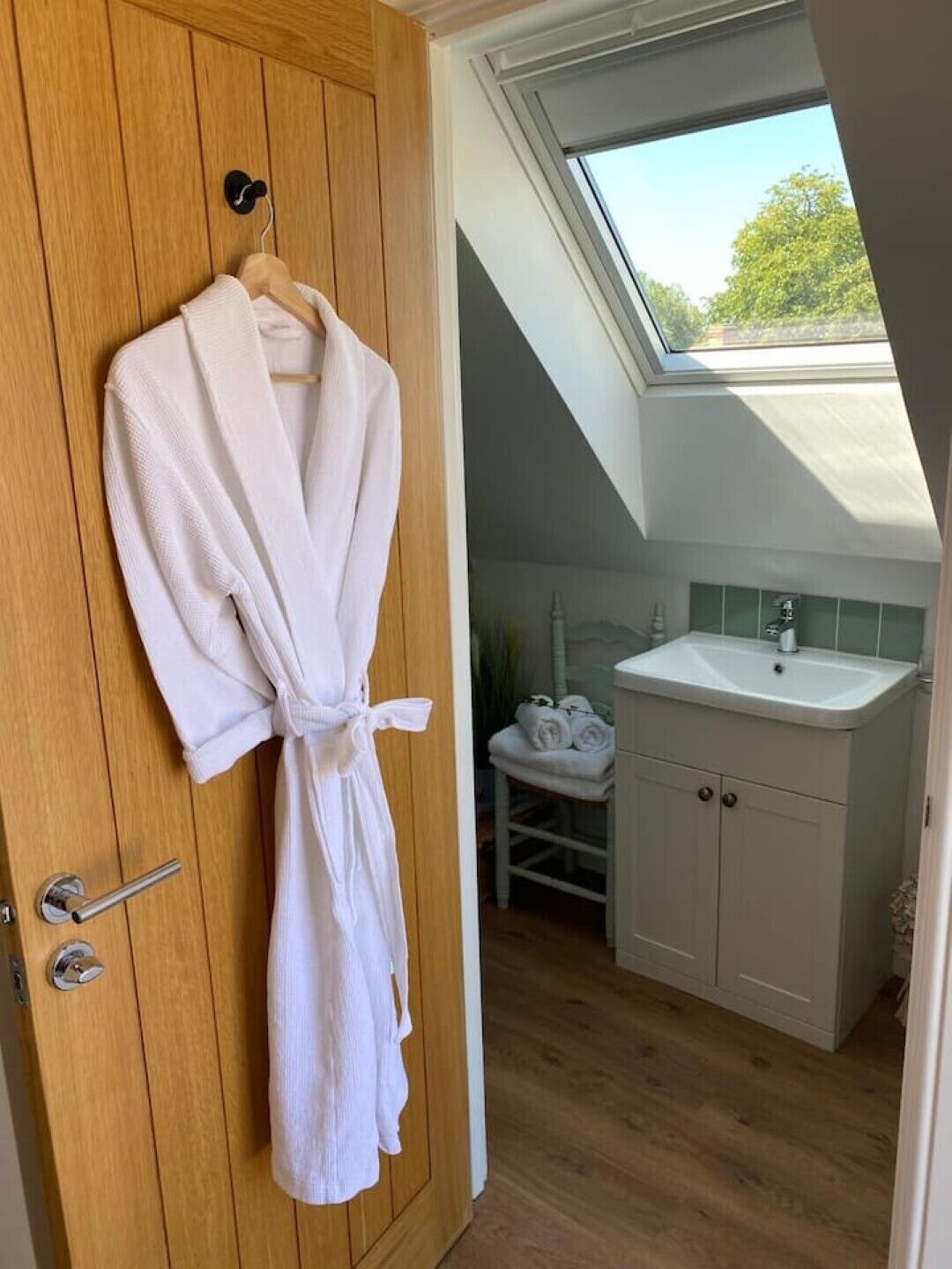 A room with a wooden door, a white robe hanging on it, a skylight, a white sink, and a small white chair.