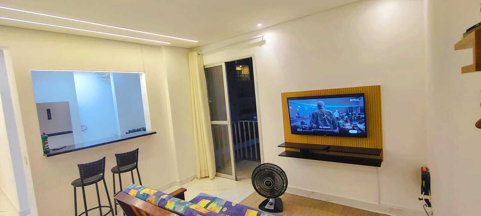 Un salón moderno con un televisor de pantalla plana montado en la pared, una mesita con dos sillas, un ventilador de pie y un ventanal corredero que da a un balcón.