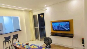 A modern living room with a flat-screen TV mounted on the wall, a small table with two chairs, a standing fan, and a sliding glass door leading to a balcony. - Apartment with pool, just a 9-minute walk from the beach! (Guarujá)