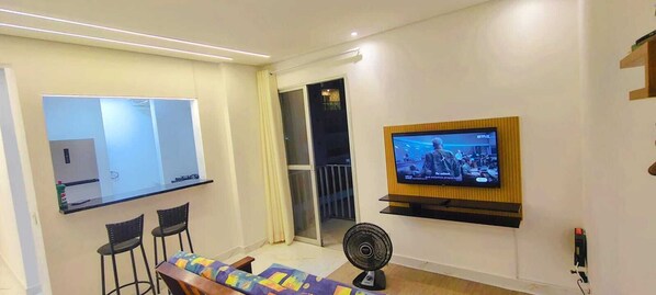 A modern living room with a flat-screen TV mounted on the wall, a small table with two chairs, a standing fan, and a sliding glass door leading to a balcony.