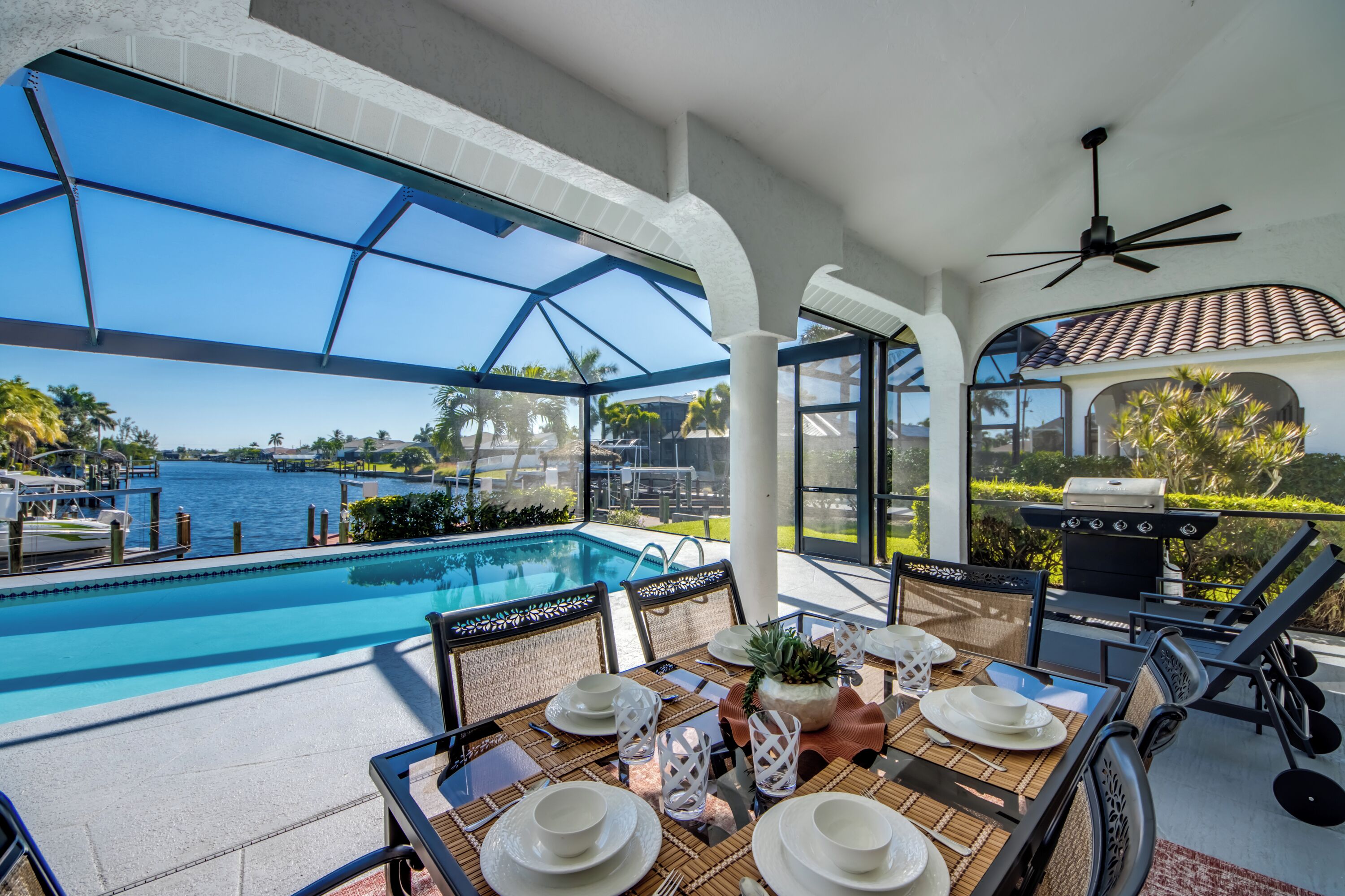 A poolside dining area with a view of the water, outdoor seating, and a grill.