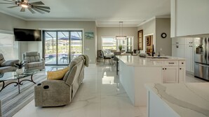 A spacious living area with a sofa, coffee table, and a ceiling fan. A kitchen with a large island and stainless steel appliances is visible. - Villa Sunset Retreat - Modern 4 BR/ 2 bath waterfront Villa w. heated pool / boat dock/ direct access (Cape Coral)