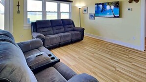 Un salon comprenant un canapé d’angle, un téléviseur à écran plat et une fenêtre équipée de stores.