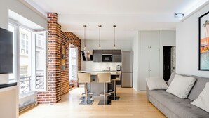 A modern living room with a grey sofa, a TV, and a kitchen area with a brick wall, bar stools, and pendant lights. - Chic apartment in the heart of Montmartre Parisian charm & central calm (Paris)