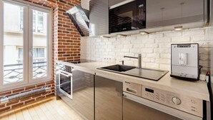 A modern kitchen with a brick wall, white subway tiles, stainless steel appliances, and a window overlooking a balcony. - Chic apartment in the heart of Montmartre Parisian charm & central calm (Paris)