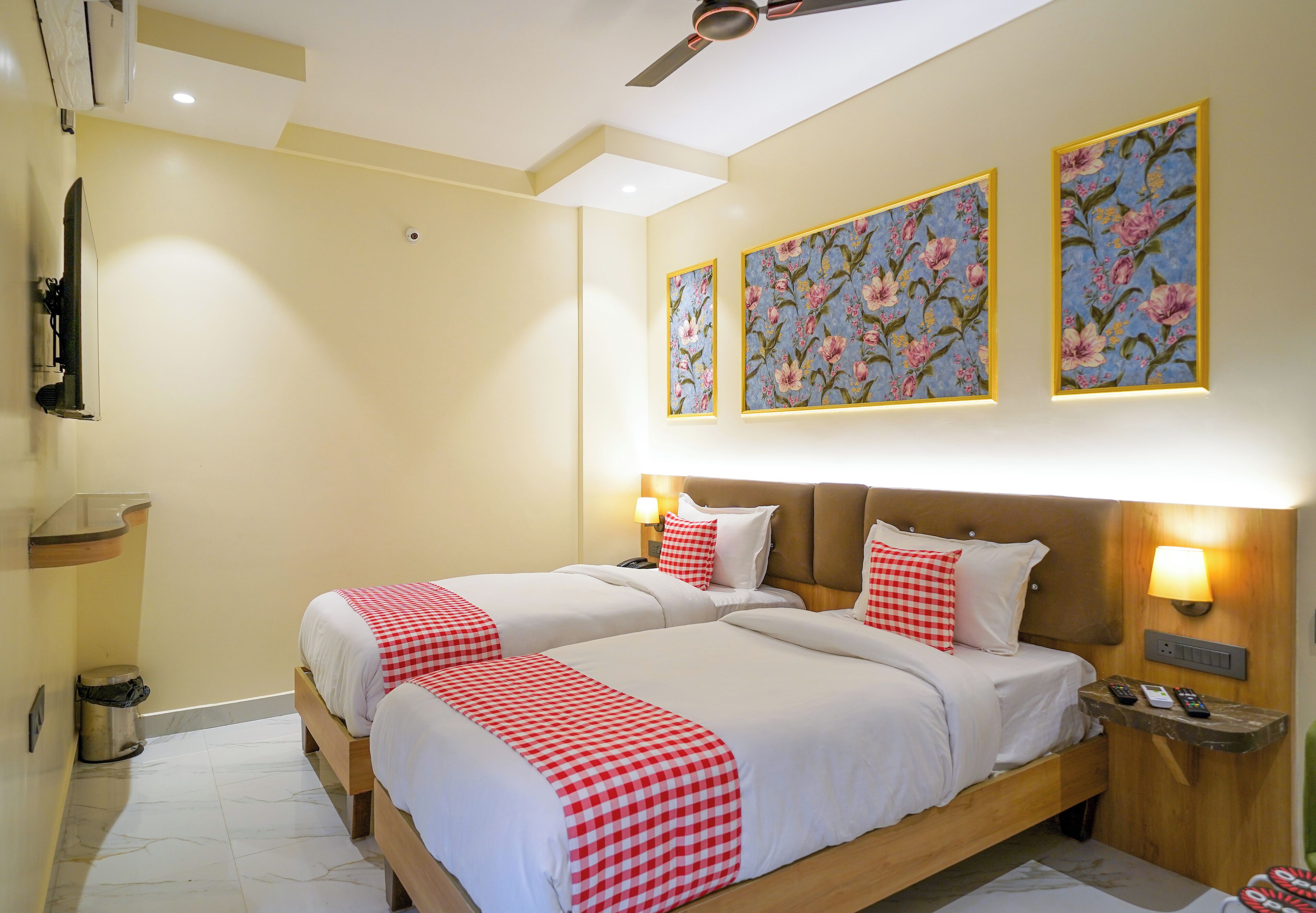 Habitación de hotel con dos camas, ventilador de techo, televisor y cuadros con motivos florales en las paredes.