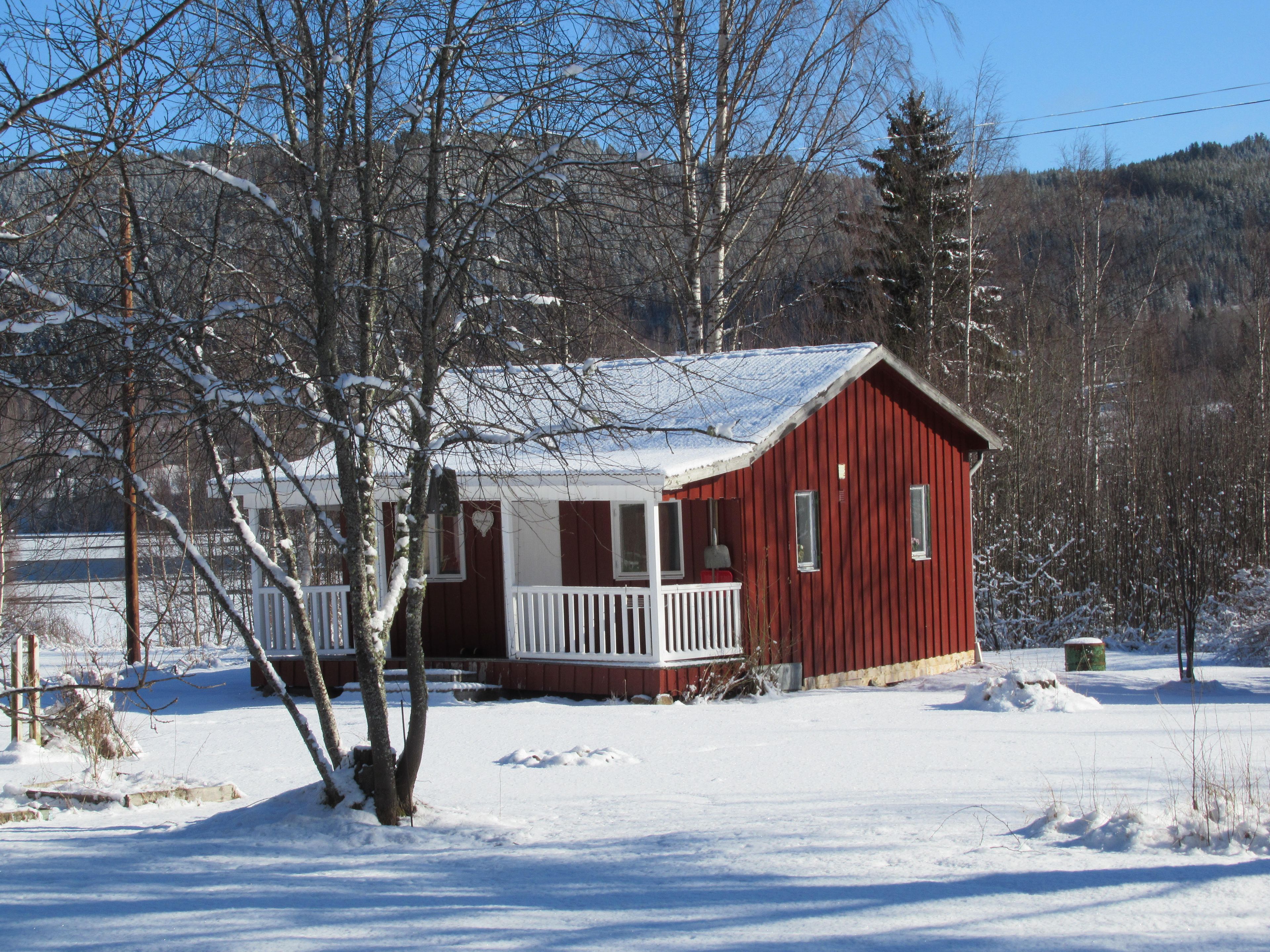 A red cabin with a white porch, surrounded by snow-covered ground and trees.