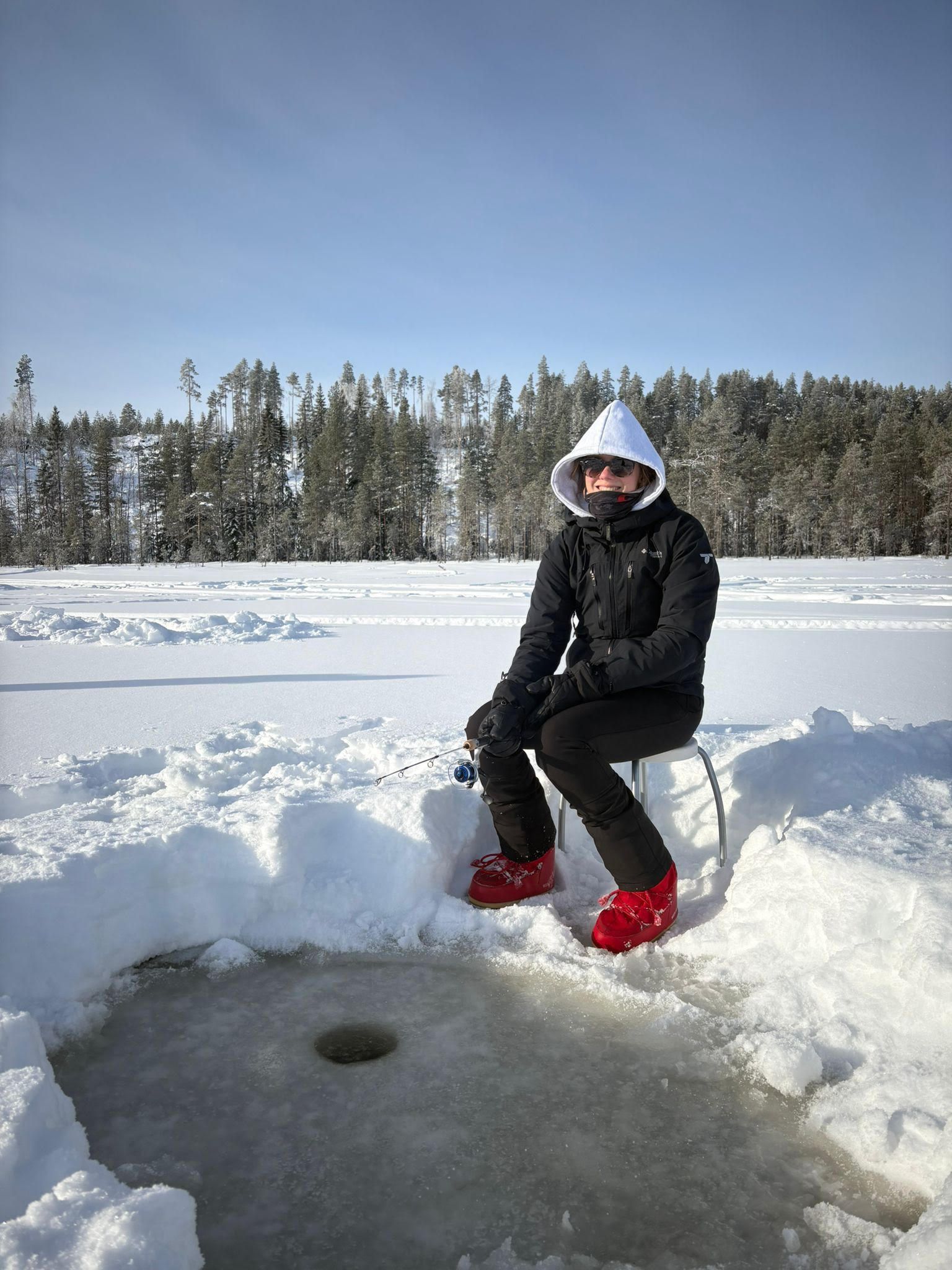 Una persona sentada en una silla en un entorno nevado al aire libre, con un agujero en el hielo, rodeada de árboles.