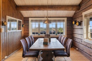 A wooden cabin interior with a dining table, chairs, a hanging light fixture, and large windows offering a view. - Trysilfjell 615 by Vacation Trysil (Trysil)