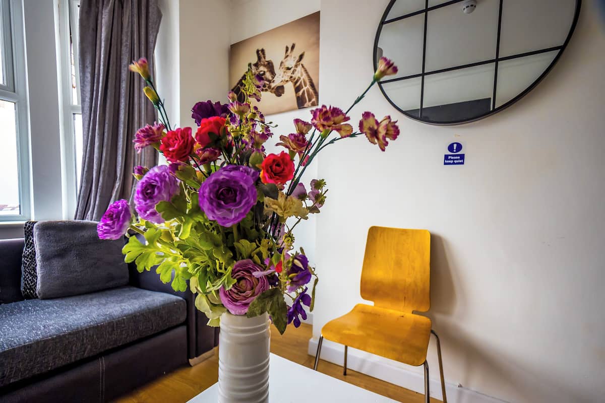 Een kamer met een witte tafel, waarop een witte vaas met bloemen staat, een grijze bank, een gele stoel, en een schilderij van giraffen aan de muur.