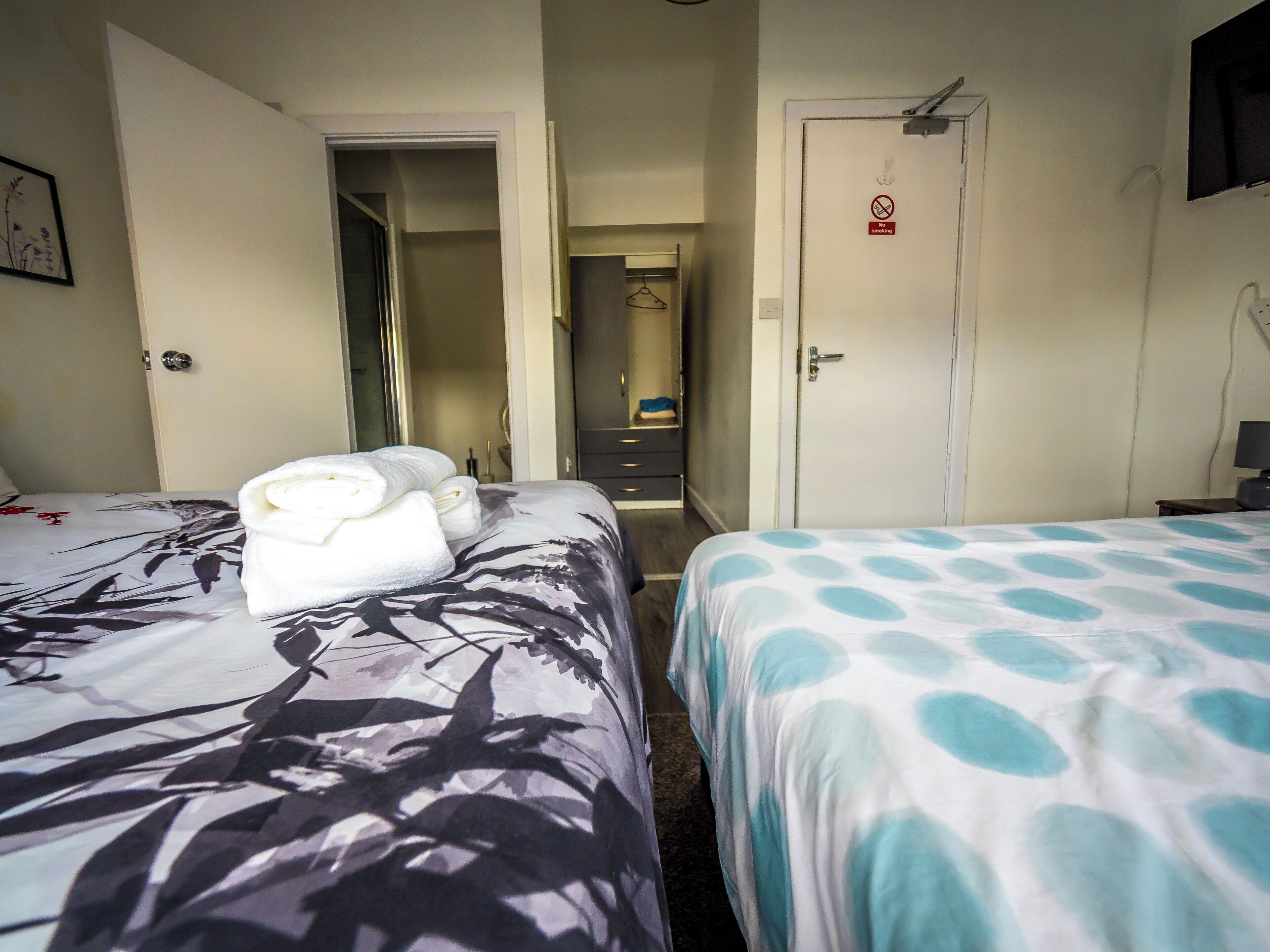 A hotel room with two beds, one with a floral bedspread and the other with a polka dot bedspread. There is a door with a 'No Smoking' sign, a mirror, and a small table with a lamp.