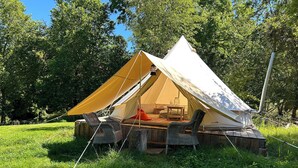 Une tente dotée d’une terrasse en bois et de chaises en osier, installée dans un espace verdoyant avec des arbres en arrière-plan.