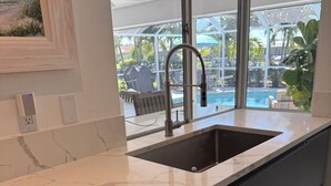 A modern kitchen with a large sink, marble countertop, and a view of a pool and outdoor seating area. - 3/2 Luxury Waterfront Pool Home (Punta Gorda)