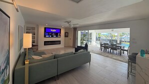 A modern living room with a large flat-screen TV, a sofa, a floor lamp, and a view of an outdoor pool area. - 3/2 Luxury Waterfront Pool Home (Punta Gorda)