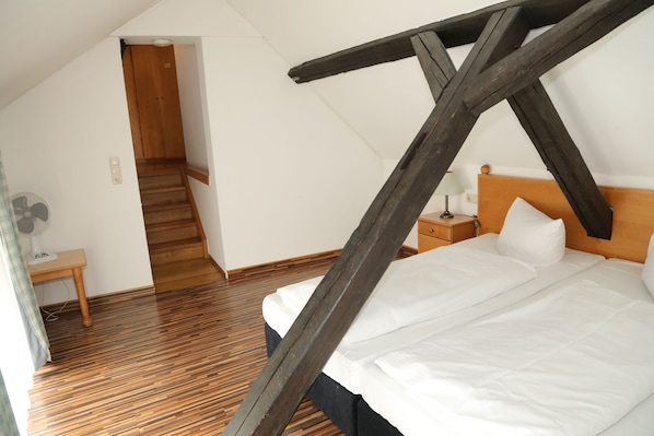 Un dormitorio con techo inclinado, suelo de madera, una cama con ropa de cama blanca, una mesita de noche y una lámpara.