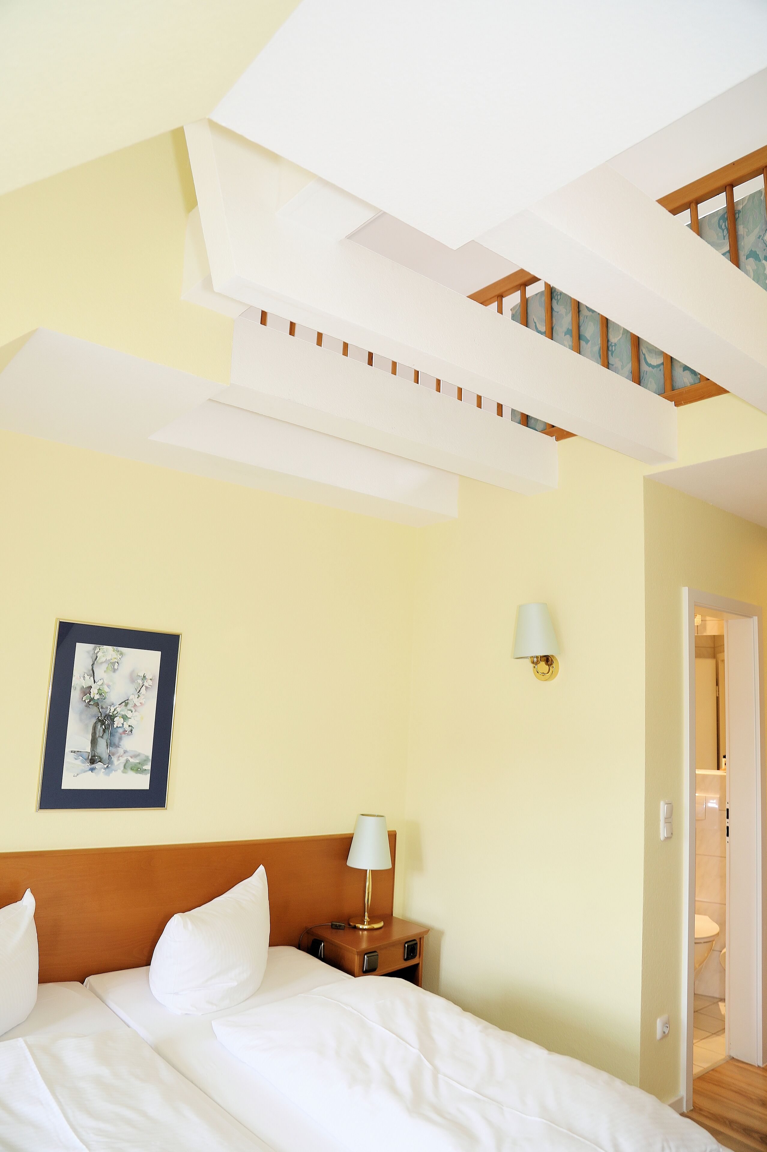 A hotel room with a bed, a nightstand, a lamp, a framed picture on the wall, and a staircase leading to an upper level.