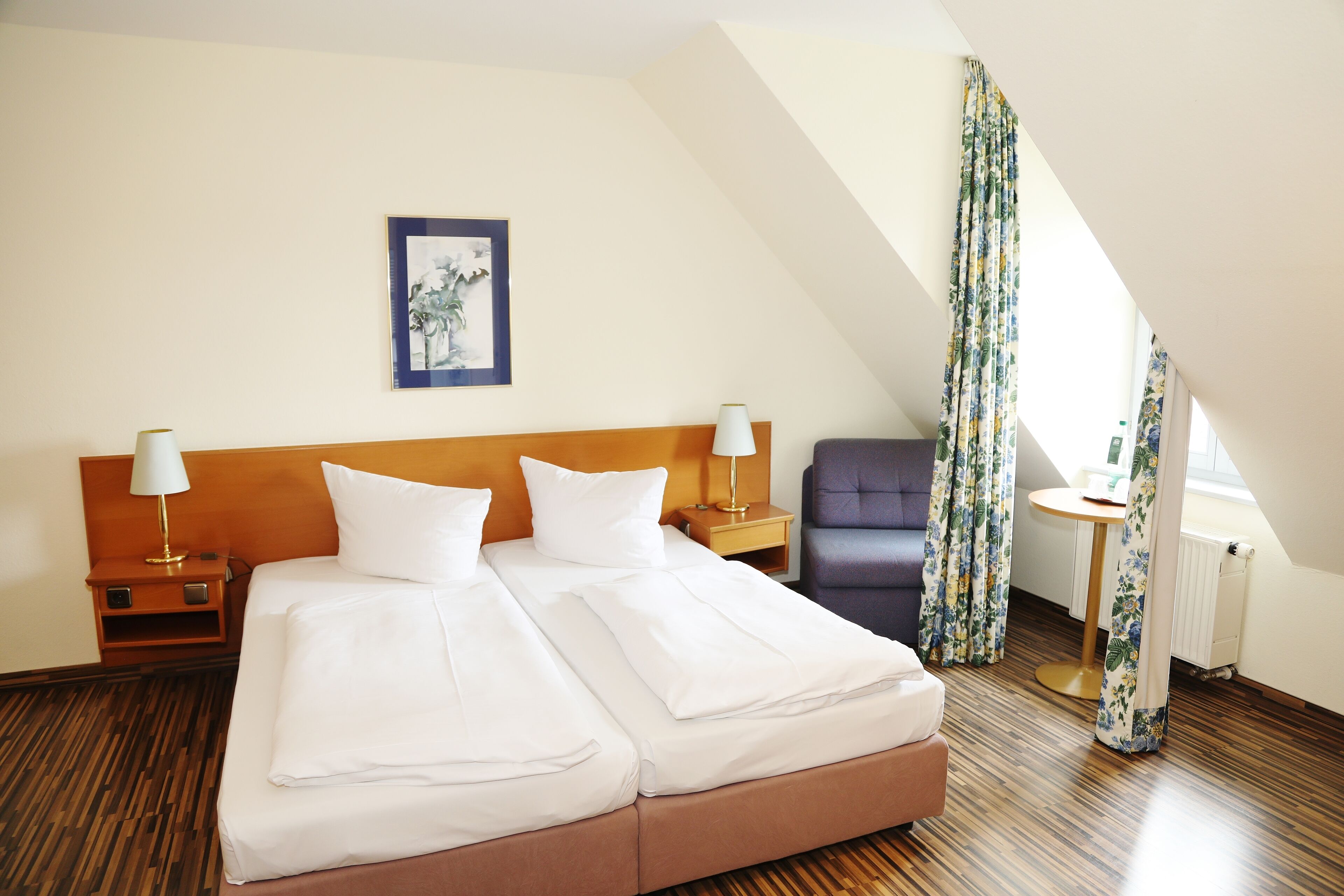 A hotel room with a large bed, two bedside tables with lamps, a chair, a small round table, and a window with curtains.