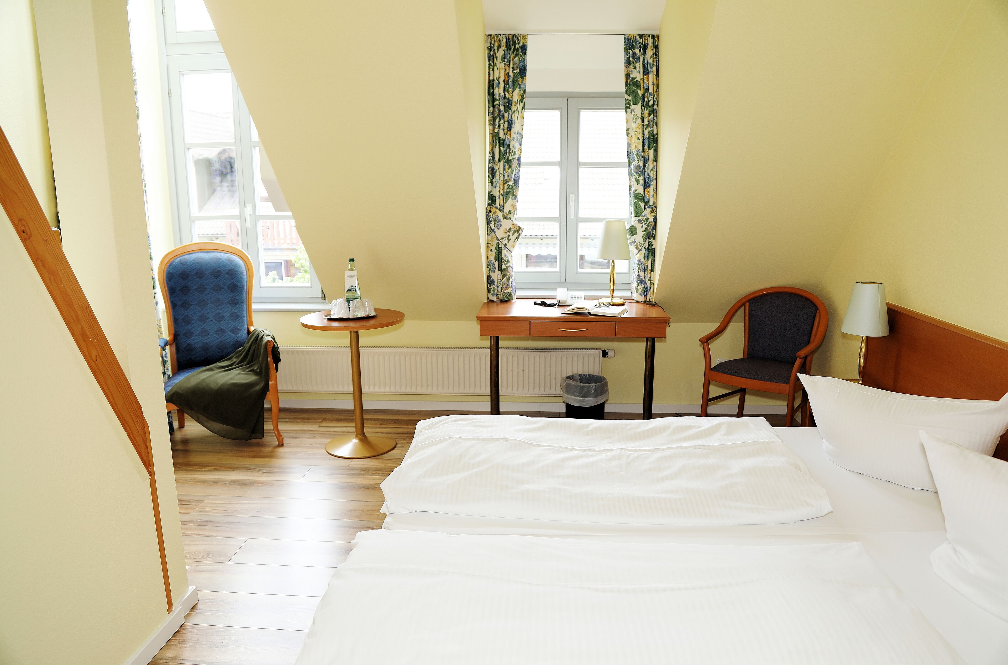 Ein Hotelzimmer mit einem Bett, zwei Stühlen, einem kleinen Tisch mit einer Lampe und einem Fenster mit Vorhängen.