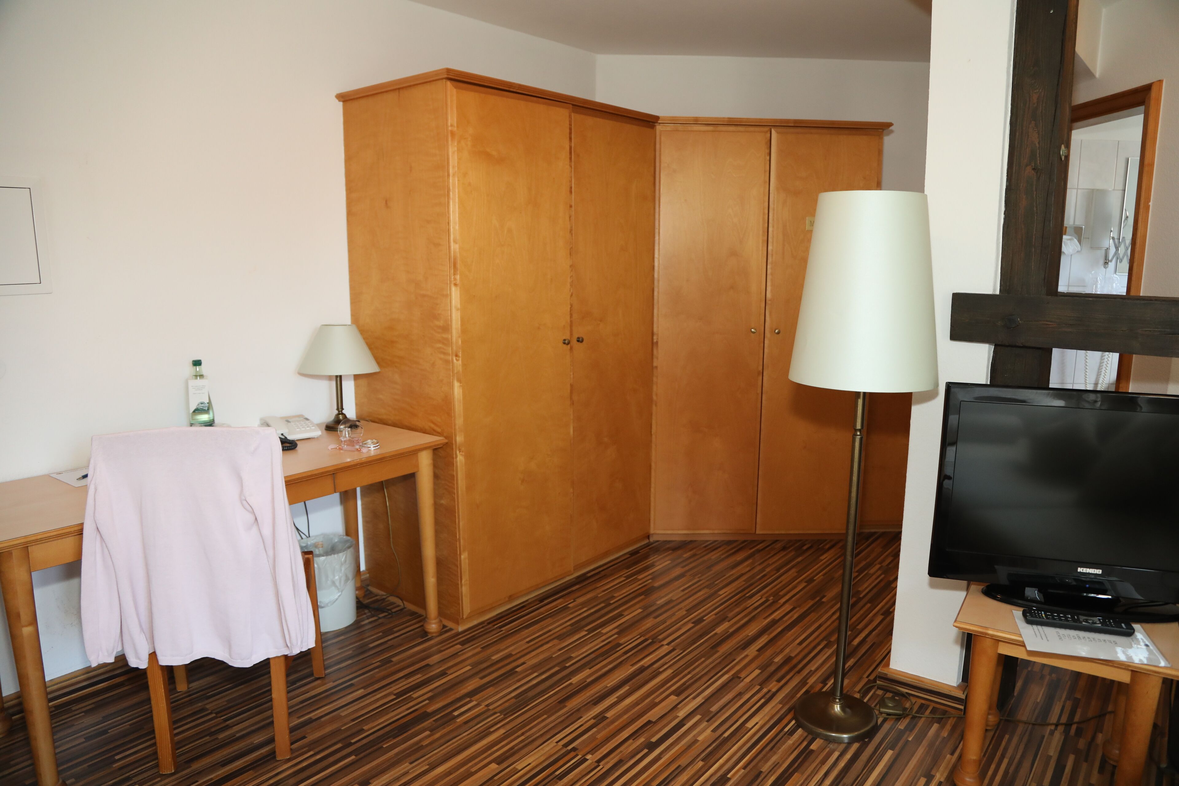 A hotel room with a wooden wardrobe, a desk with a lamp, a television, and a floor lamp.