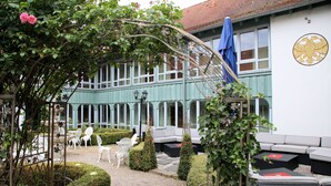 Ein Innenhof mit einer weißen Außenlounge, einer mit Kletterpflanzen bewachsenen Pergola und einem Gebäude mit blauem Markisenvorzelt und einem Adlerwappen.