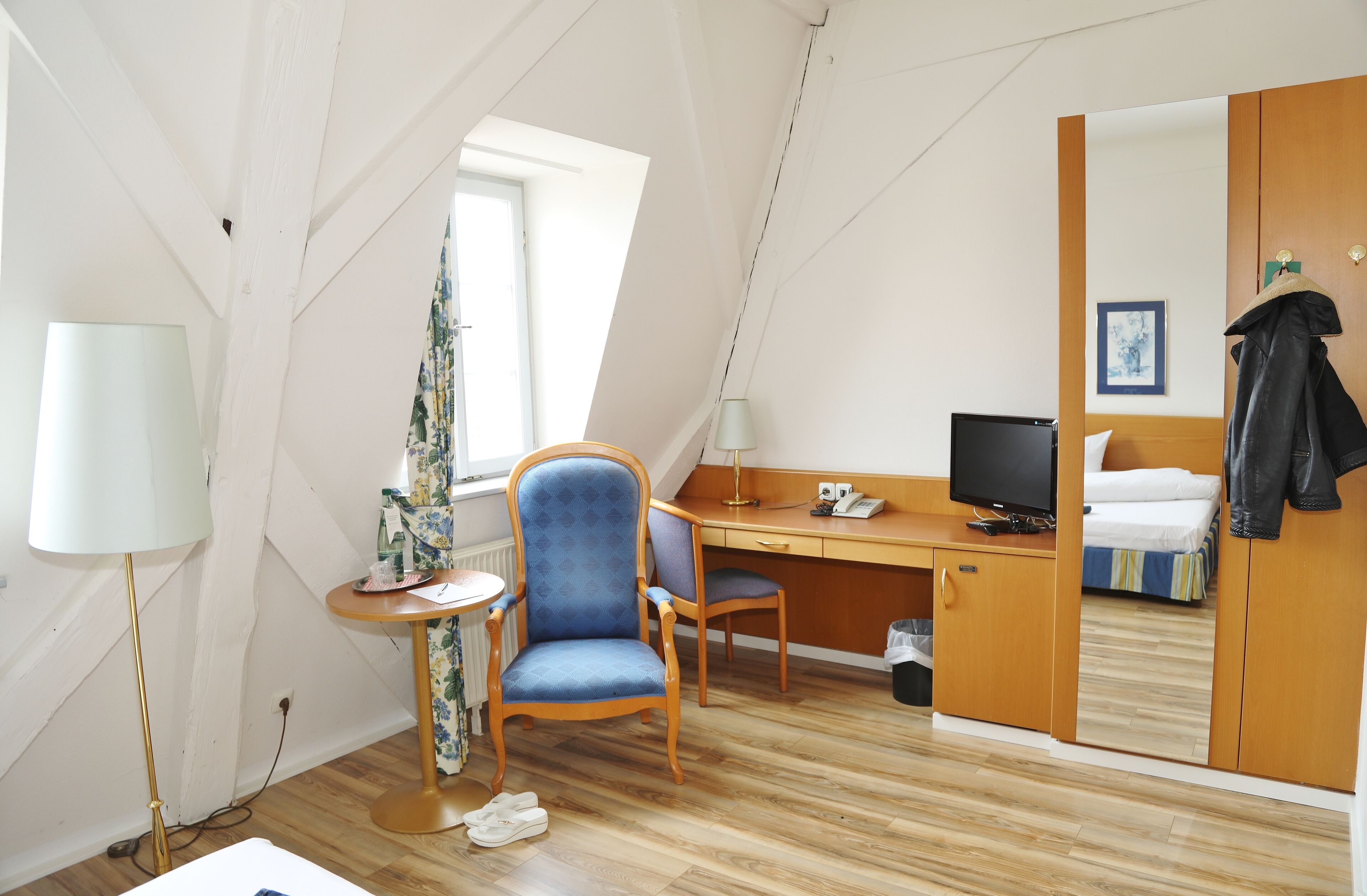 Una habitación con escritorio de madera, una silla azul, una cama, un televisor y un espejo.