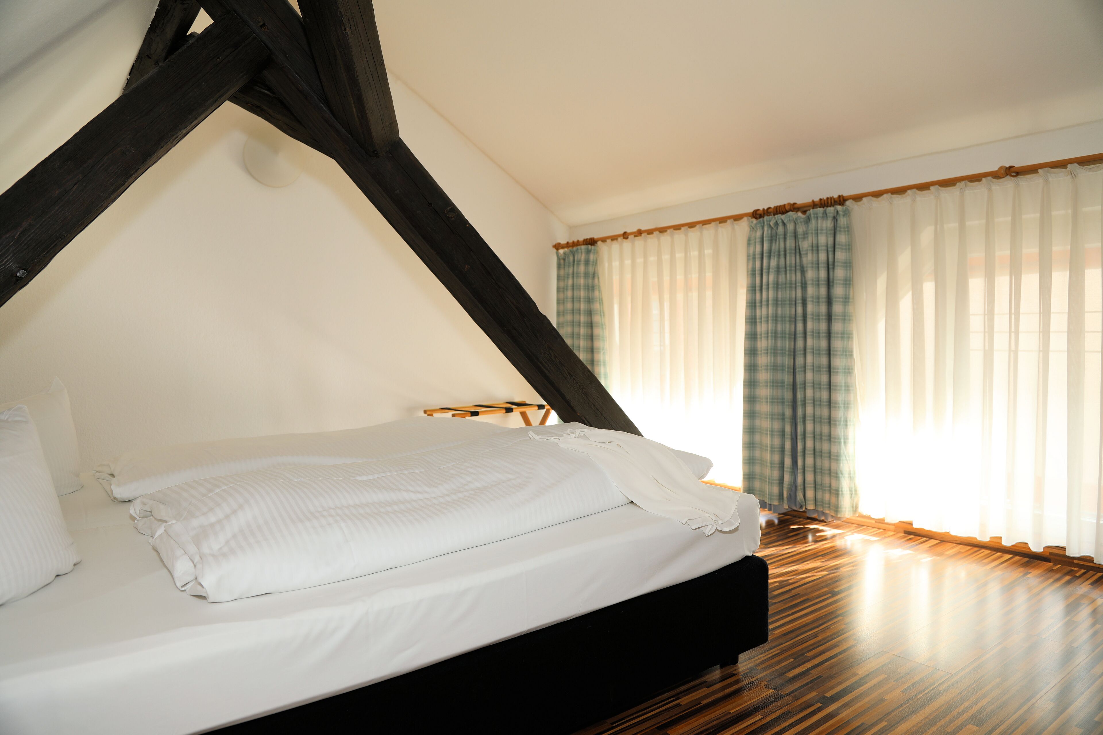 A bedroom with a bed, wooden beams, white curtains, and a wooden floor.