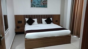 A hotel room with a large bed, a nightstand, a painting on the wall, and a window with curtains. - Gokul Dham Haridwar (Haridwar)
