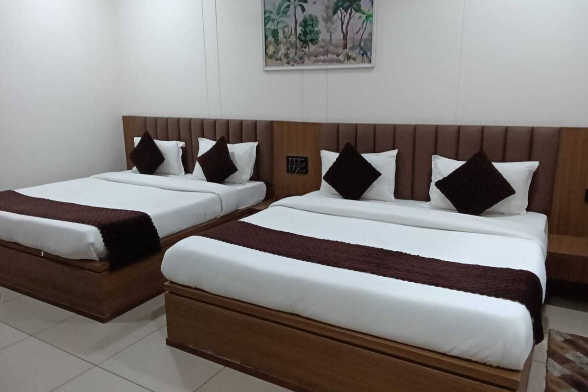 A hotel room with two single beds, each with a wooden headboard, white bedding, and dark throw pillows. A painting of a tropical scene hangs on the wall above the beds.