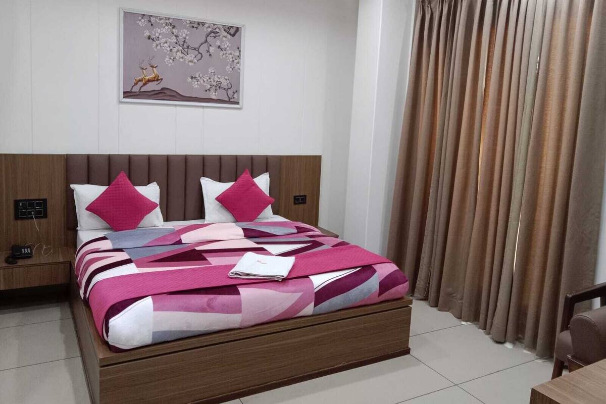 A modern hotel room with a large bed, a headboard, a nightstand, a wall-mounted power outlet, and a window with curtains.