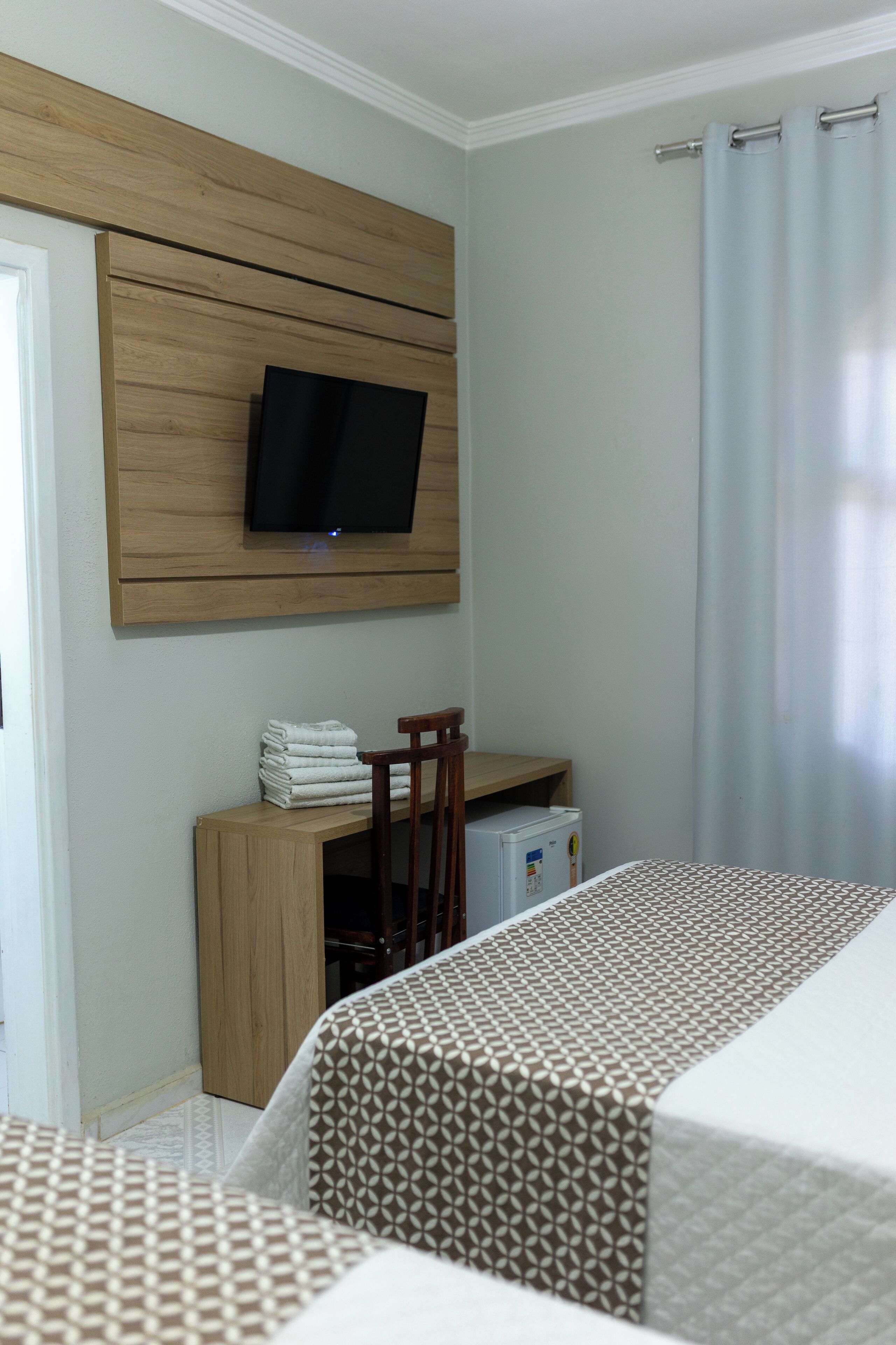 Een hotelkamer met een flatscreen-tv bevestigd aan een wand van houten panelen, een klein houten bureau met een stoel, een koelkast en twee bedden met beddengoed met een patroon.