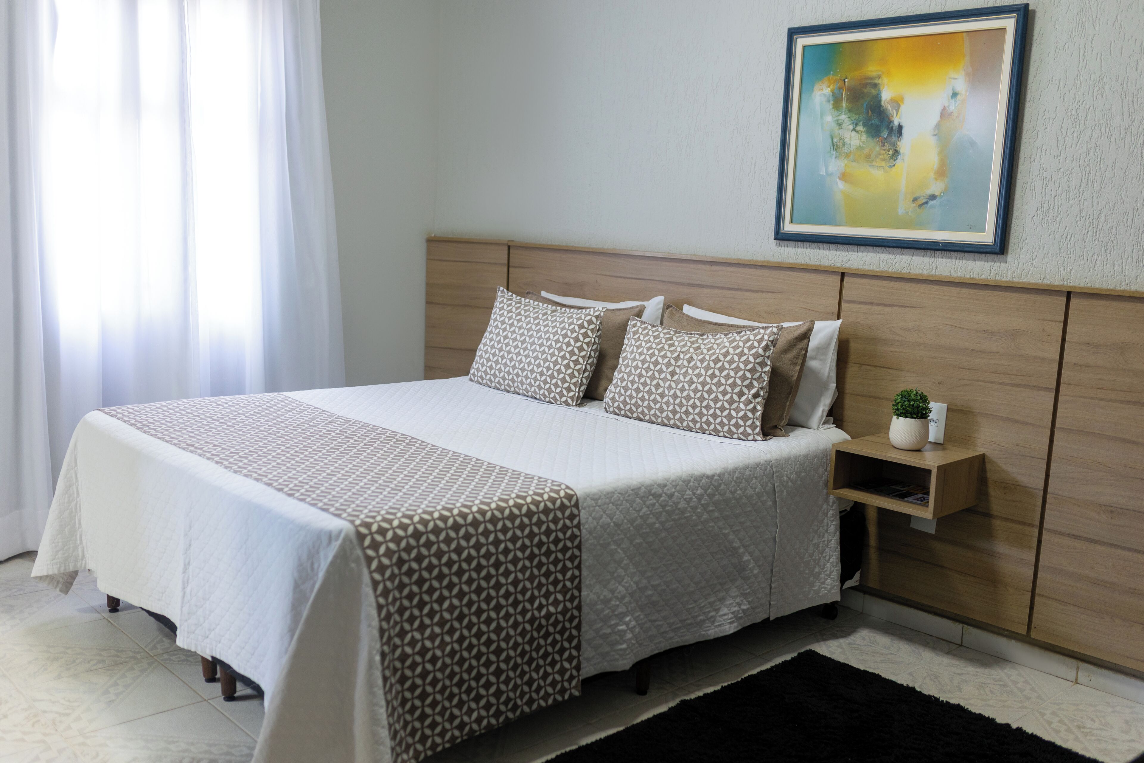 A neatly made bed with a patterned bedspread, a bedside table with a plant, and a framed abstract painting on the wall.